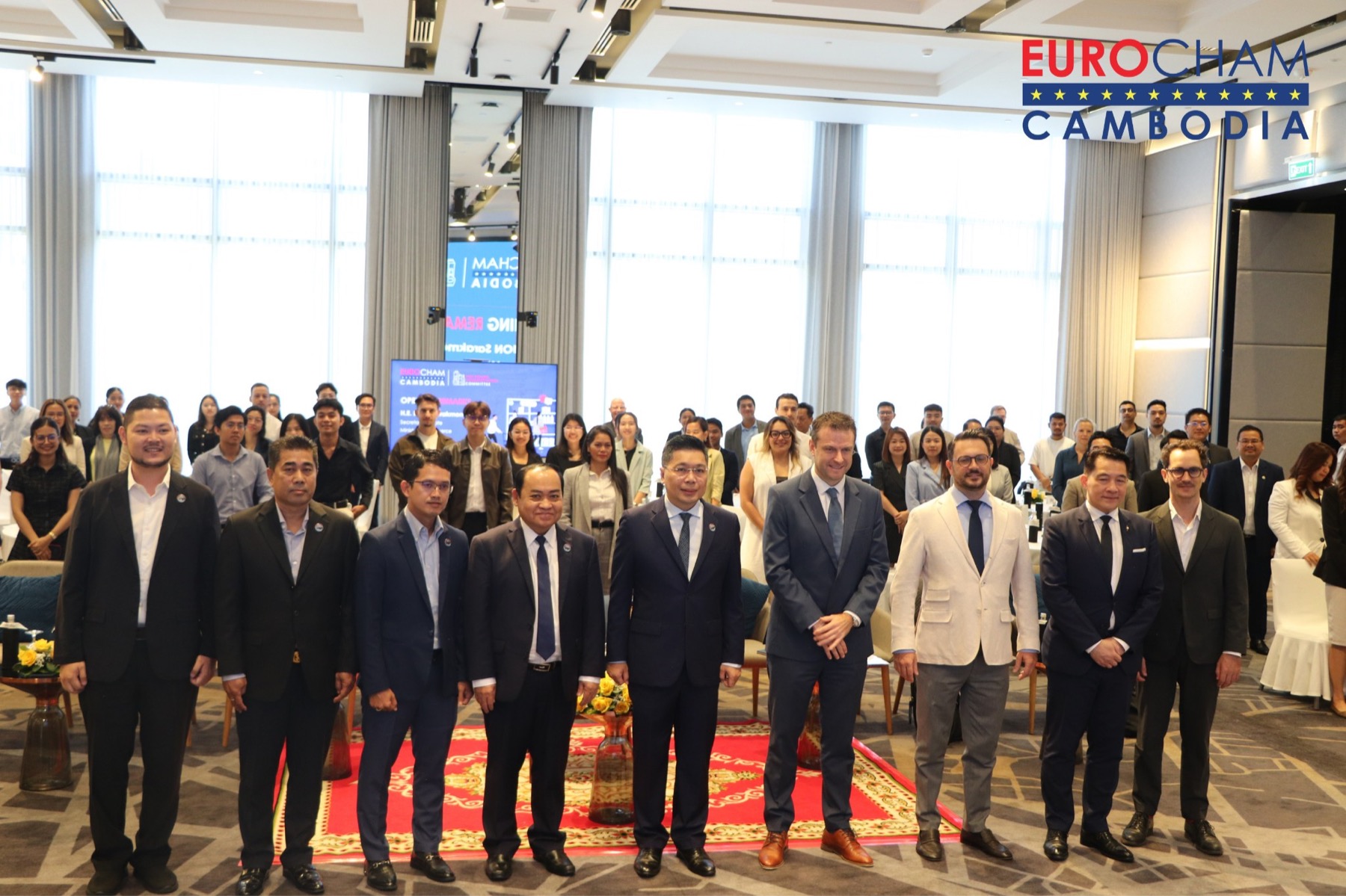 A line of guests and officials photographed in front of a room of members at a EuroCham Cambodia event